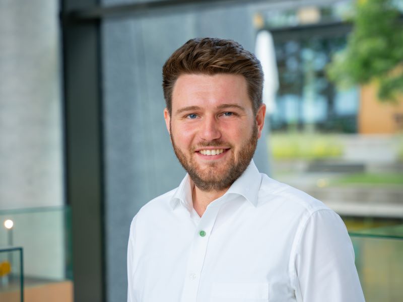 Eine männliche Person mit weißem Hemd steht im Foyer eines Firmengebäudes.